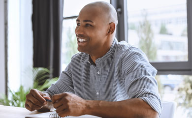 A smiling young professional male wearing a button down shirt with a tablet of paper and pen in front of him