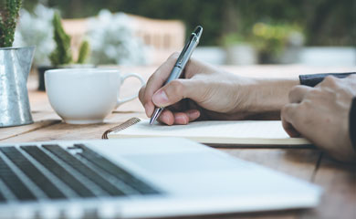 Hand holding pen writing on tablet of paper with laptop in the foreground